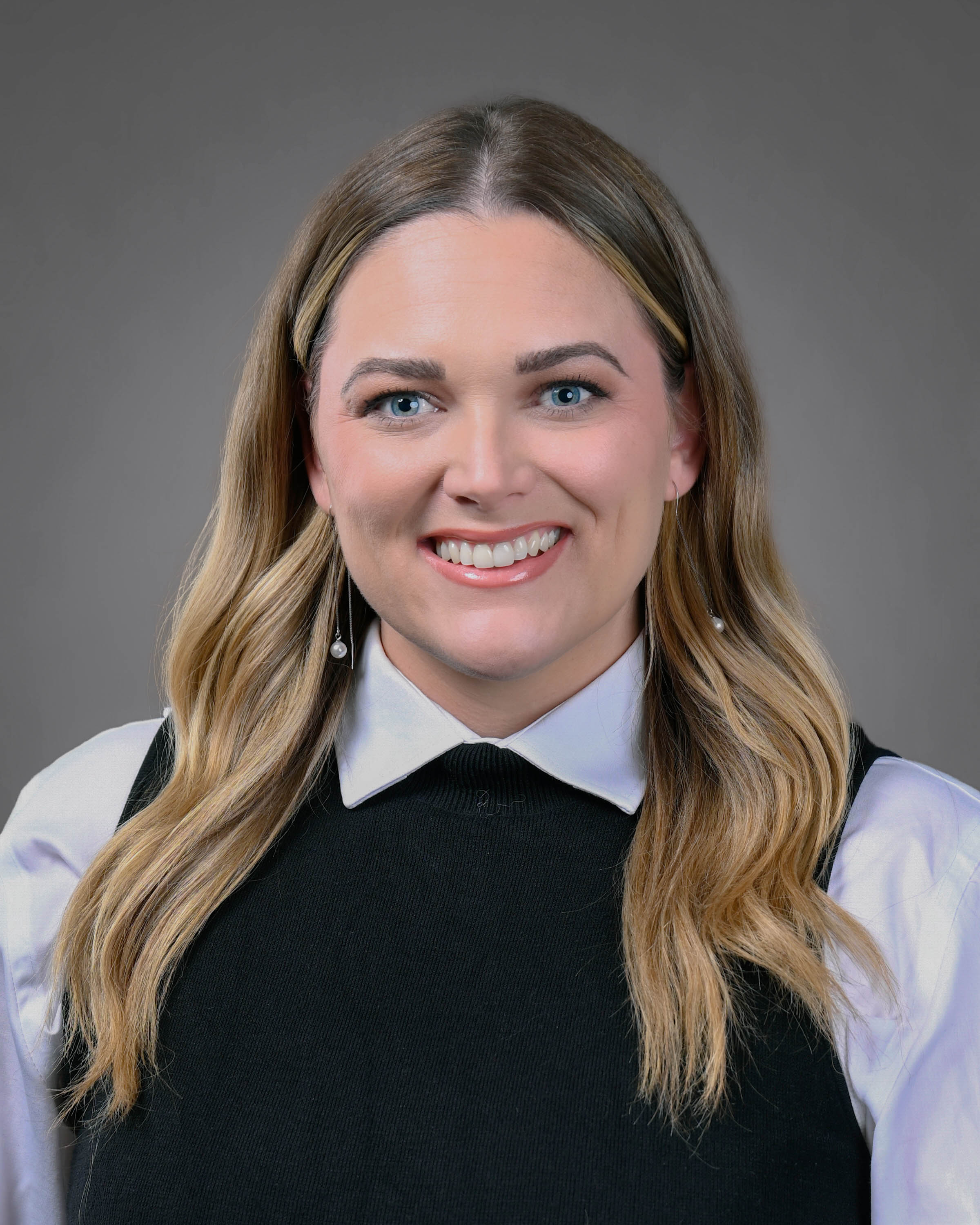Professional headshot of a smiling woman with long wavy blonde hair, wearing a black sleeveless top over a white collared blouse against a gray studio background.